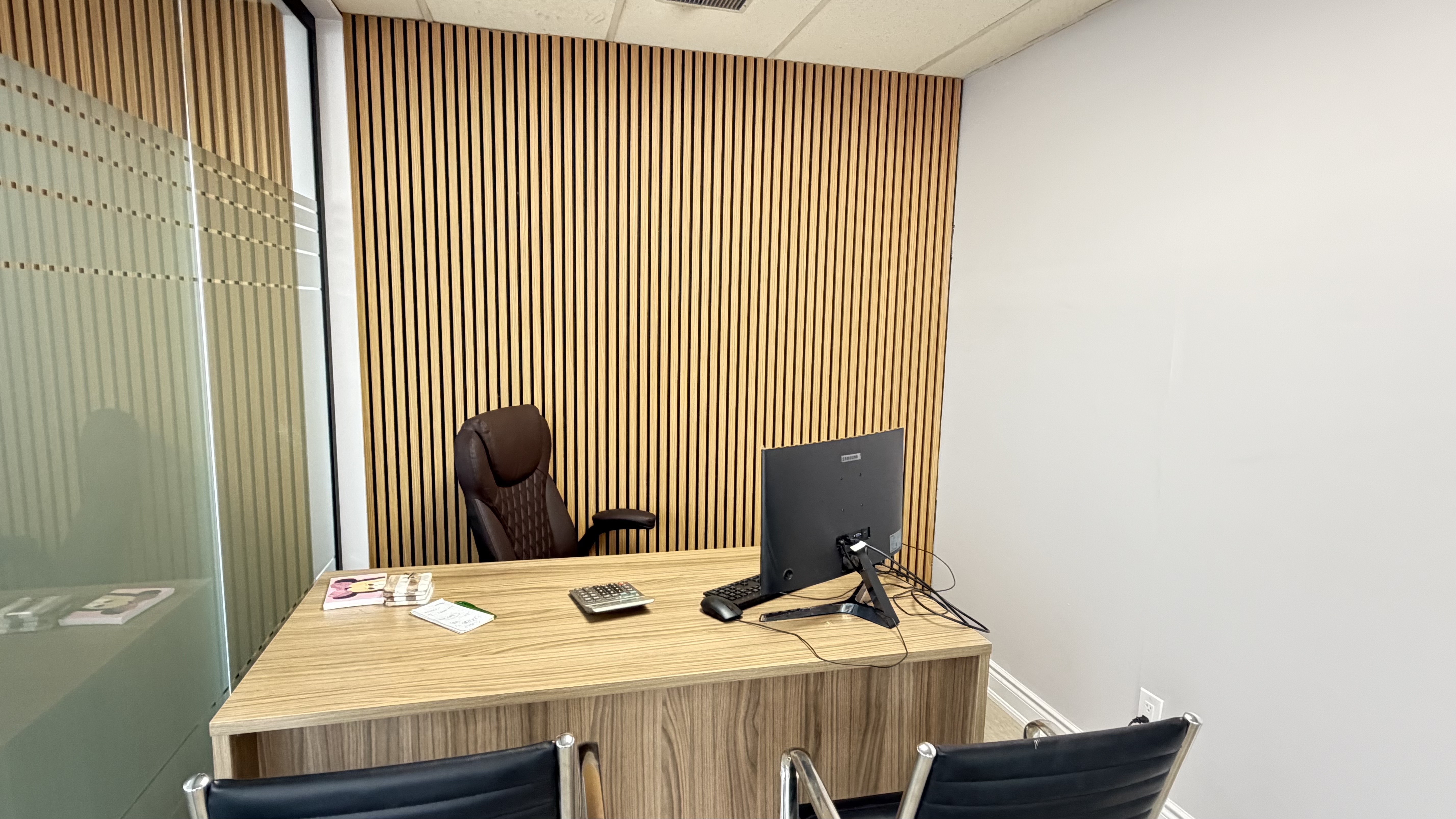 A modern private office featuring a wooden desk with a computer monitor, a brown leather executive chair, black guest chairs, and a contemporary vertical wood slat accent wall next to a glass partition